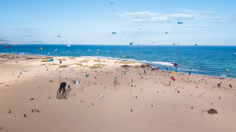 Fuerteventura Menschen lassen zahlreiche bunte Drachen über einem weiten Sandstrand am Meer steigen. Am Himmel fliegen Figuren in Tier- und Fantasieformen. Im Hintergrund das blaue Meer mit sanften Wellen.