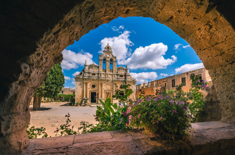 Blick durch einen Steinbogen auf ein Klostergebäude in Arkadi mit Glockenturm, Innenhof und Blumen im Vordergrund