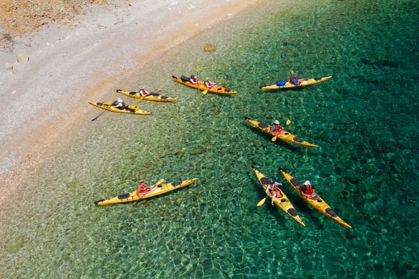 Kroatien Gruppe von Menschen in gelben Kajaks auf kristallklarem, türkisfarbenem Wasser in der Nähe eines steinigen Strandes. Die Paddler tragen Schwimmwesten und Sonnenhüte.