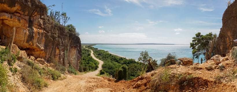 Panoramablick von einem felsigen Küstenpfad auf eine tropische Landschaft mit grüner Vegetation, Sandweg und türkisfarbenem Meer