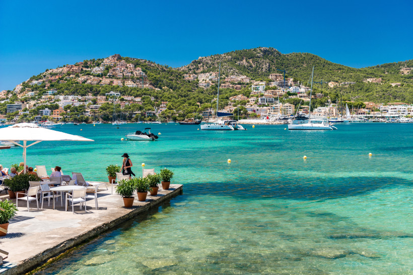 Mallorca Sonnige Küstenlandschaft mit kristallklarem türkisfarbenem Wasser. Im Vordergrund eine Terrasse mit weißen Tischen und Stühlen, auf der Menschen unter weißen Sonnenschirmen sitzen. Im Hintergrund grüne Hügel mit vielen Häusern, sowie mehrere Segelboote un