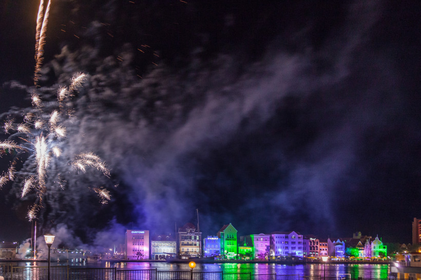 Curaçao Nächtliches Feuerwerk über der bunt beleuchteten Häuserzeile von Willemstad, Curaçao. Links steigen silberne Raketen in den Himmel, während sich am Wasser bunte Lichter spiegeln. Dichte Rauchschwaden ziehen über die Szenerie.