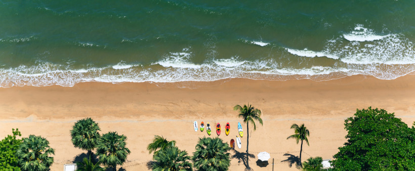 Thailand Pattaya Luftaufnahme eines tropischen Strandes mit Palmen, bunten Kajaks und Stand-up-Paddle-Boards im Sand. Kleine Wellen treffen auf den goldenen Sand, das Wasser schimmert grünlich-blau