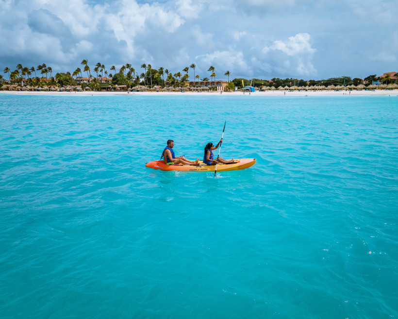 Zwei Personen paddeln gemeinsam in einem orange-gelben Kajak auf türkisblauem, ruhigem Meer. Im Hintergrund ein heller Sandstrand mit Palmen und einigen Hotelgebäuden. Der Himmel ist leicht bewölkt.