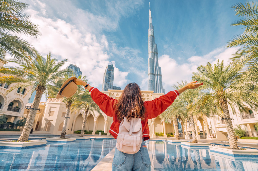 Dubai Eine junge Frau mit Rucksack steht vor einem Palmenhof und blickt mit ausgestreckten Armen auf den Burj Khalifa in Dubai, den höchsten Wolkenkratzer der Welt. Sie trägt eine rote Jacke und hält einen Sonnenhut in der Hand. Der Himmel ist blau mit einigen