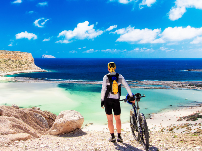 Kreta Ein Radfahrer steht mit seinem Mountainbike auf einer Anhöhe und blickt auf die türkisfarbene Balos-Lagune auf Kreta. Vor ihm breitet sich eine atemberaubende Küstenlandschaft mit Sandstränden und tiefblauem Meer aus. Der Himmel ist strahlend blau mit ver