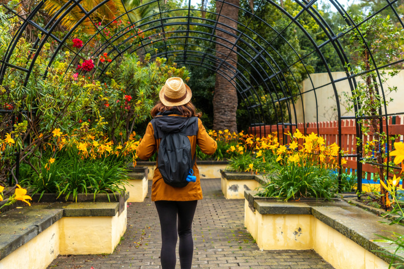 Botanischer Garten in Arucas – Natur erleben beim Spaziergang Frau mit Rucksack spaziert durch einen blühenden botanischen Garten in Arucas. Farbenfrohe Blumen, tropische Pflanzen und ein Rankbogen sorgen für mediterranes Urlaubsfeeling.