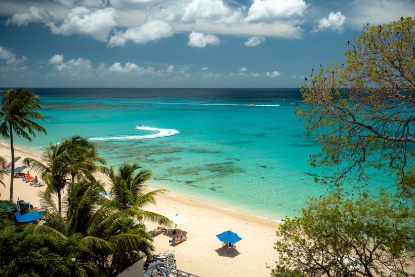 Barbados Blick auf einen weißen Sandstrand auf Barbados mit türkisfarbenem Meer. Palmen säumen den Strand, im Wasser zieht ein Jetski eine weiße Spur. Einige Sonnenliegen mit blauen Schirmen stehen im Schatten der Bäume, der Himmel ist teils bewölkt.