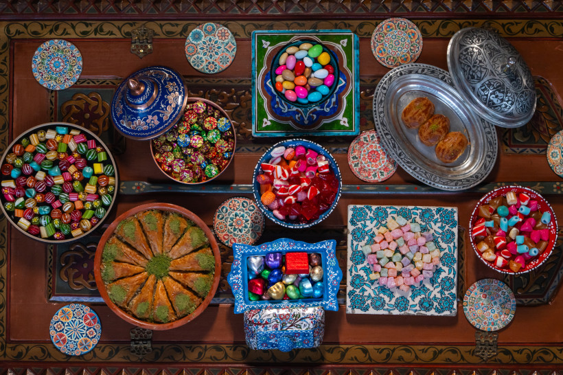 Verschiedene bunte Süßigkeiten und traditionelle Desserts sind kunstvoll in Schalen auf einem orientalisch verzierten Tisch angerichtet. Darunter Baklava, gefüllte Bonbons, Lokum und Pralinen – alles in festlicher Atmosphäre.