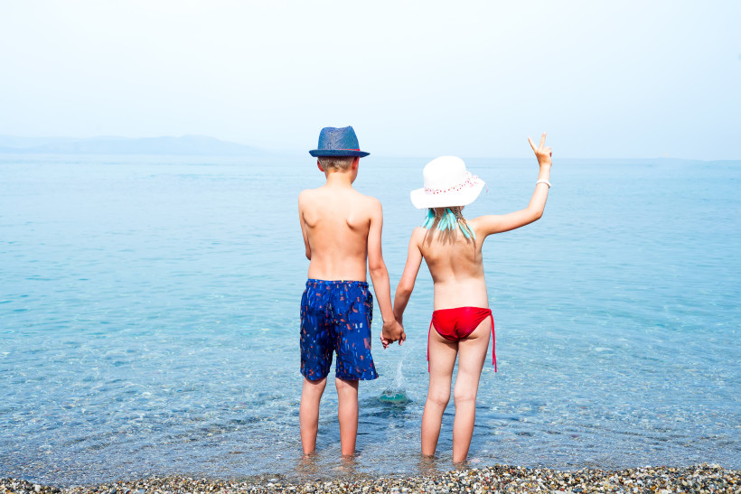 Zwei Kinder stehen Hand in Hand im seichten Wasser am Kiesstrand und blicken aufs Meer hinaus. Das Kind links trägt eine blaue Badehose und einen blauen Hut, das Kind rechts einen weißen Sonnenhut und einen roten Bikini. Es hebt die freie Hand und zeigt e