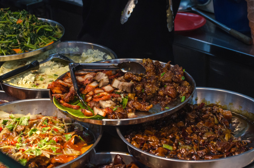 Singapur Nahaufnahme einer Auswahl an Streetfood in Singapur. In mehreren Edelstahlschalen befinden sich gebratene Fleischstücke, würzige Soßen, gebratenes Gemüse und aromatische asiatische Gerichte. Ein Koch im Hintergrund bedient das Buffet mit einer Zange