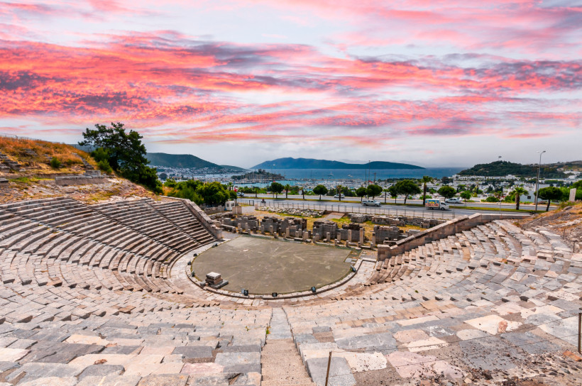 Antikes Amphitheater in Bodrum, Türkei, mit halbkreisförmigen Steinstufen und Blick auf die Stadt, das Meer und bewaldete Hügel. Der Himmel leuchtet in dramatischem Rosa und Orange bei Sonnenuntergang.