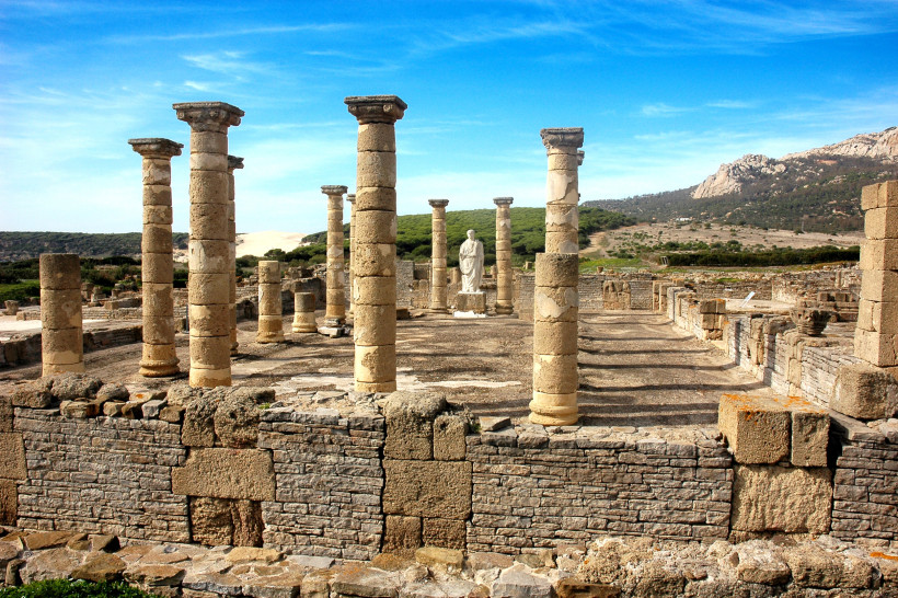 Römische Ruinen von Baelo Claudia nahe der Playa de Bolonia. Mehrere erhaltene Steinsäulen stehen in Reih und Glied, im Hintergrund eine weiße Statue, dahinter Dünen und grüne Hügel unter blauem Himmel.
