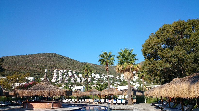 Poolanlage eines Hotels mit Palmen, Strohdach-Sonnenschirmen und Blick auf weiße Häuser am Hang