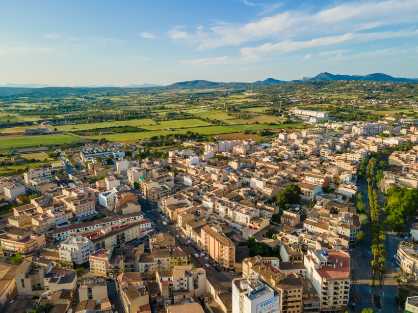 Mallorca - Manacor Luftaufnahme einer mittelgroßen Stadt mit vielen flachen Gebäuden in warmen Erdtönen, umgeben von weiten, grünen Feldern und Hügeln am Horizont. Die Stadt ist von kleinen Straßen durchzogen, einige davon sind von Palmen gesäumt. Im Vordergrund sieht man e
