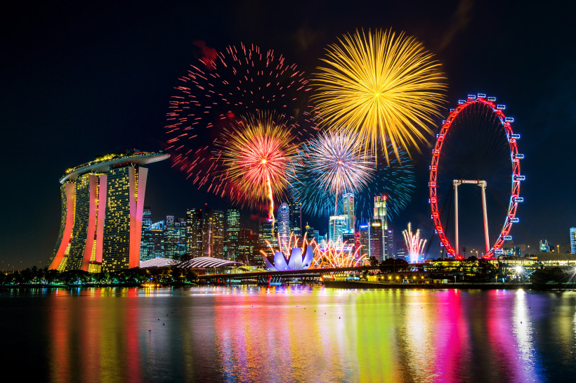 Singapur Farbenfrohes Feuerwerk über der Marina Bay in Singapur. Zu sehen sind das markante Hotel Marina Bay Sands, das rot beleuchtete Riesenrad Singapore Flyer und zahlreiche Hochhäuser der Skyline. Die Spiegelung der Lichter im Wasser rundet die nächtliche Szen