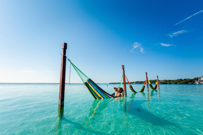Mexiko Mann entspannt in einer bunten Hängematte über türkisblauem Wasser, befestigt an Holzpfosten, unter einem klaren blauen Himmel