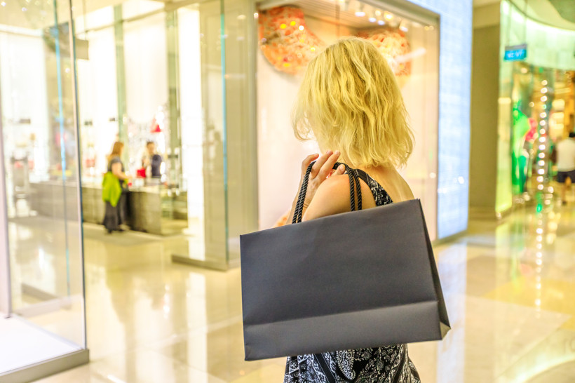 Singapur Blonde Frau von hinten in einem modernen Einkaufszentrum. Sie trägt ein Sommerkleid und eine große schwarze Einkaufstasche über der Schulter und schaut in ein hell beleuchtetes Schaufenster.