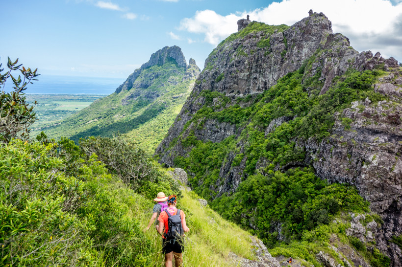 Mauritius Dieses Bild zeigt eine abenteuerliche Wanderung durch eine atemberaubende Berglandschaft. Zwei Wanderer, ausgestattet mit Rucksäcken und Hüten, bewegen sich vorsichtig auf einem schmalen, grasbewachsenen Pfad entlang eines steilen Hangs. Die Berge sind ei