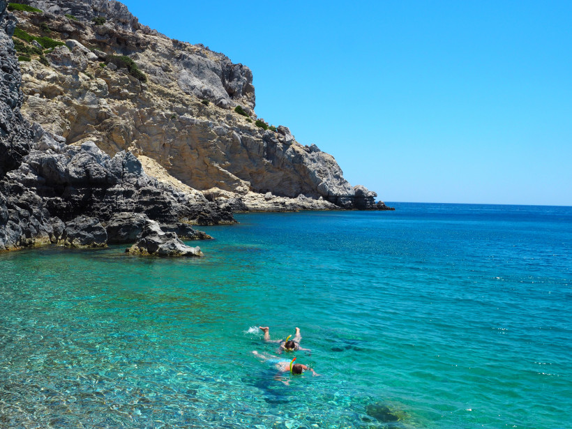 Das Bild zeigt eine kleine, abgelegene Badebucht mit kristallklarem Wasser in verschiedenen Türkistönen.   Links und im Hintergrund ragen steile, teils grau-schwarze Felswände in die Höhe, teilweise mit beigem Gestein durchzogen.   Im Vordergrund schnorch