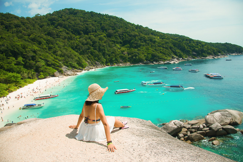 Thailand Eine Frau mit Sonnenhut, Bikini und weißem Rock sitzt auf einem Felsen oberhalb einer tropischen Bucht mit türkisfarbenem Wasser. Sie blickt auf einen weißen Sandstrand, an dem viele Menschen baden und entspannen. Im Wasser ankern mehrere Schnellboote und