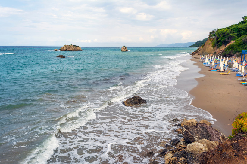 Prasoudi Beach mit Sandstrand, Wellen und Felsen Blick auf den Prasoudi Beach mit Sandstrand, Meereswellen, Felsen im Wasser und Strandliegen mit Sonnenschirmen am Ufer