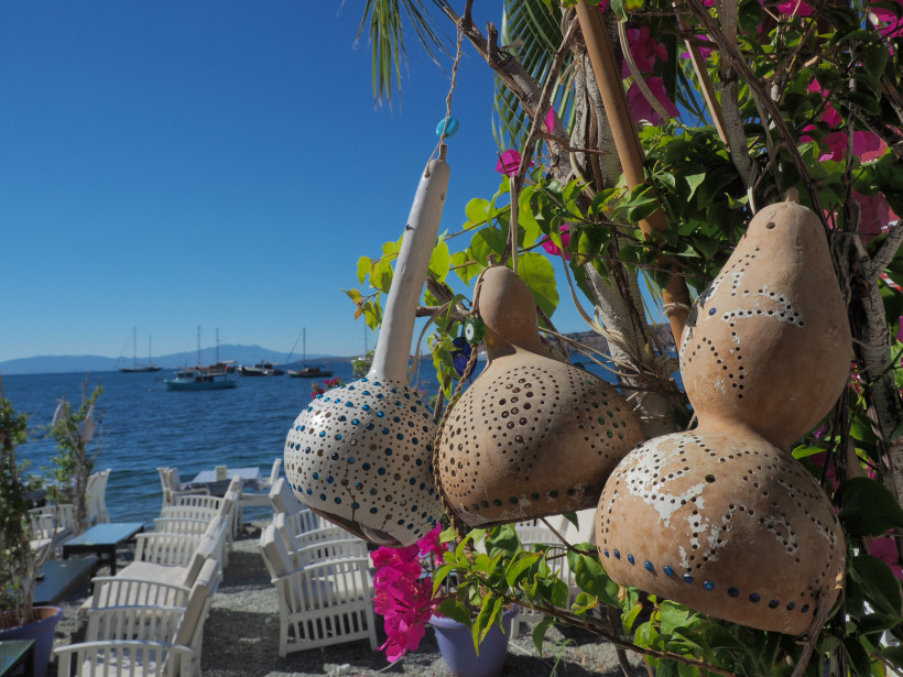 Handverzierte Trockenfrüchte-Lampen hängen an einer blühenden Pflanze vor einem kleinen Restaurant direkt am Meer. Im Hintergrund sieht man weiße Stühle, Tische und segelnde Boote auf dem ruhigen Wasser unter klarem, blauem Himmel.