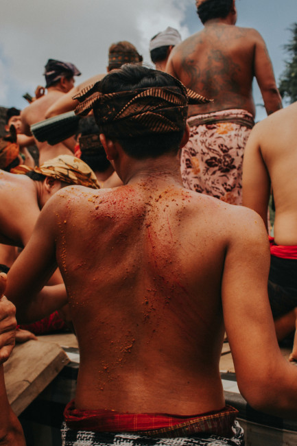 Bali Ein junger balinesischer Mann mit nacktem Oberkörper und traditioneller Kopfbedeckung ist von hinten zu sehen. Sein Rücken zeigt rote Kratzspuren und Rückstände von Kräutern oder Gewürzen, vermutlich im Rahmen eines spirituellen Rituals. Um ihn herum steh