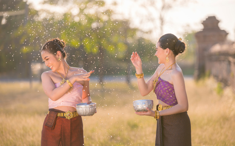 Thailand Zwei Frauen in traditioneller thailändischer Kleidung spielen lachend mit Wasser im Sonnenlicht. Beide halten verzierte Silberschalen und tragen goldenen Schmuck. Um sie herum fliegen glitzernde Wassertropfen.