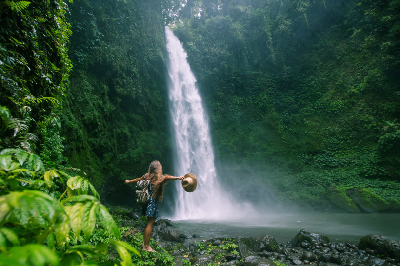 Bali Eine Frau mit langem Haar, Sommerkleid und Rucksack steht barfuß auf nassen Steinen vor einem hohen Wasserfall inmitten tropischer Vegetation auf Bali. Sie hält ihren Hut in der Hand und breitet die Arme aus, während Wassernebel die Luft durchzieht. Der W