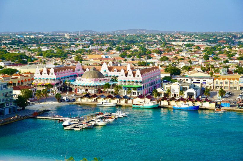 Luftaufnahme der Altstadt von Oranjestad auf Aruba. Im Vordergrund liegt der Hafen mit Booten, dahinter bunt verzierte Gebäude mit Giebeldächern und einem auffälligen runden Kuppelbau. Die Stadt erstreckt sich bis zu den Hügeln am Horizont.