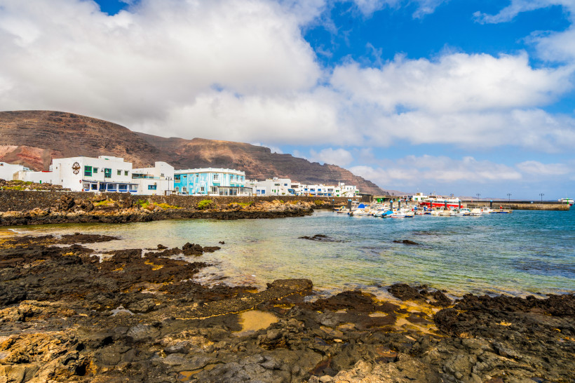 Hafen von Órzola im Norden Lanzarotes – Fischerort mit Vulkanlandschaft Der Fischerort Órzola im Norden Lanzarotes mit bunten Booten, heller Küste und vulkanischen Felsen vor eindrucksvoller Bergkulisse.