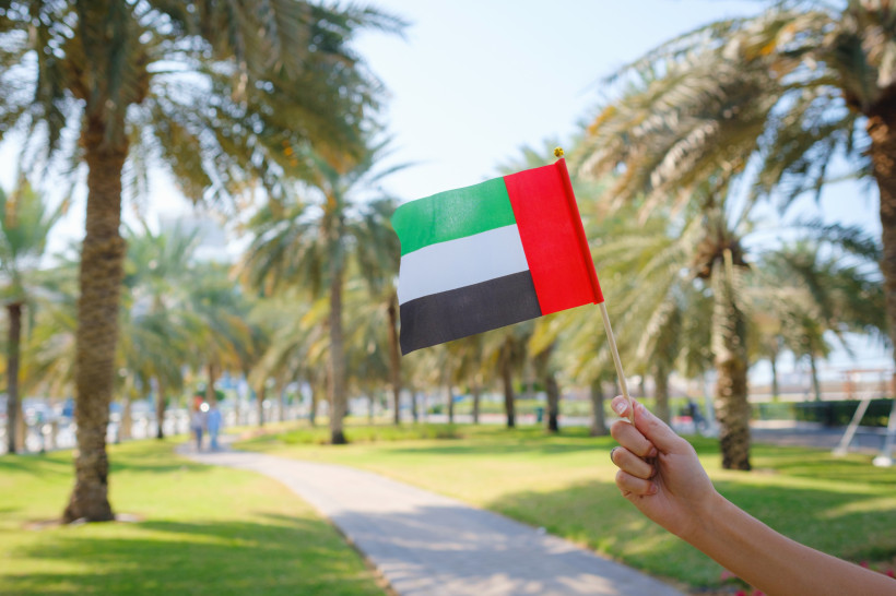 Nationalfeiertag VAE  Nahaufnahme einer weiblichen Hand, die von der Corniche aus eine kleine Flagge der Vereinigten Arabischen Emirate vor dem Abu Dhabi Park hält.