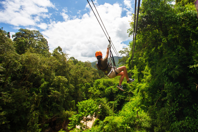 Person mit Helm und Sicherheitsgurt fährt an einer Zipline durch den tropischen Dschungel. Unten dichter grüner Wald, blauer Himmel mit Wolken im Hintergrund