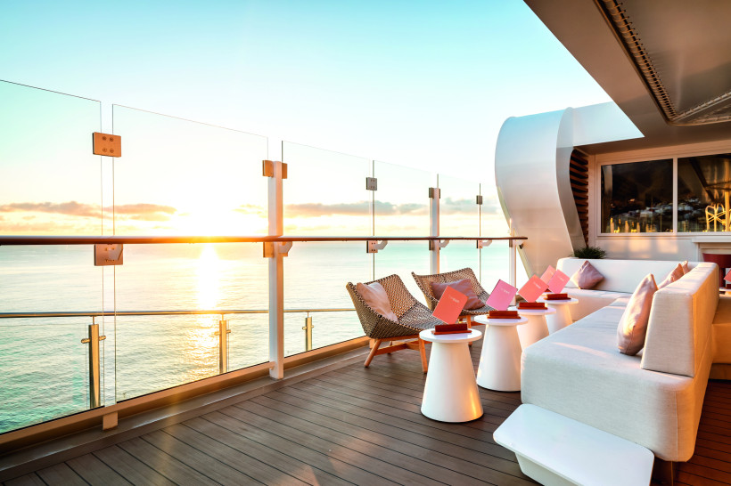 Außenbereich der Hoheluft Bar auf der Mein Schiff 1 mit Sitzgelegenheiten am Glasgeländer und Blick auf das Meer bei Sonnenuntergang