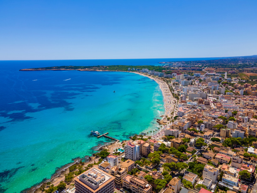 Luftaufnahme einer Küstenstadt mit langem Sandstrand, türkisblauem Meer, Hotels und Booten in einer geschwungenen Bucht