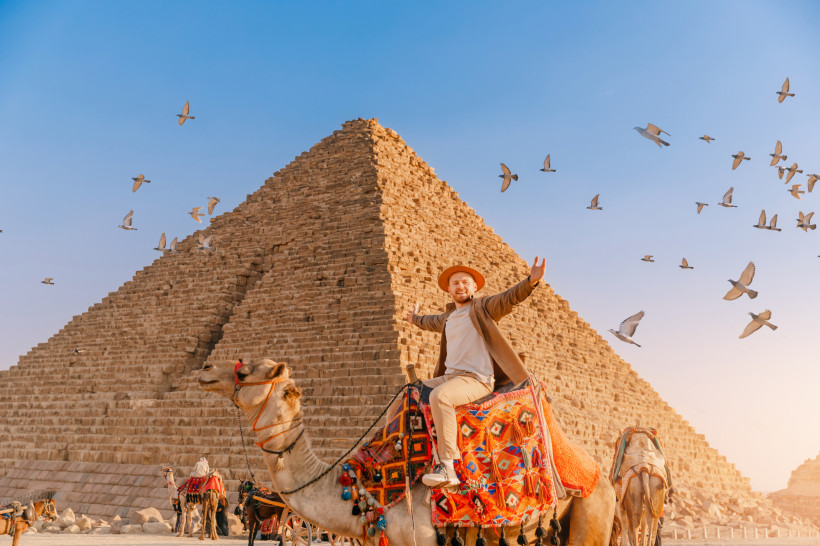 Ägypten Ein lachender Tourist sitzt auf einem bunt geschmückten Kamel vor der großen Pyramide von Gizeh. Am Himmel fliegt ein Schwarm Tauben, der Himmel ist klar und blau
