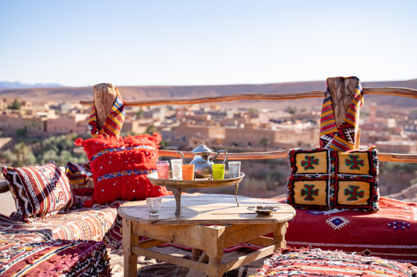 Restaurant, Marokko Orientalisch eingerichtete Terrasse mit bunten Kissen, Teetablett und Blick auf eine Wüstenstadt und Berge in der Ferne
