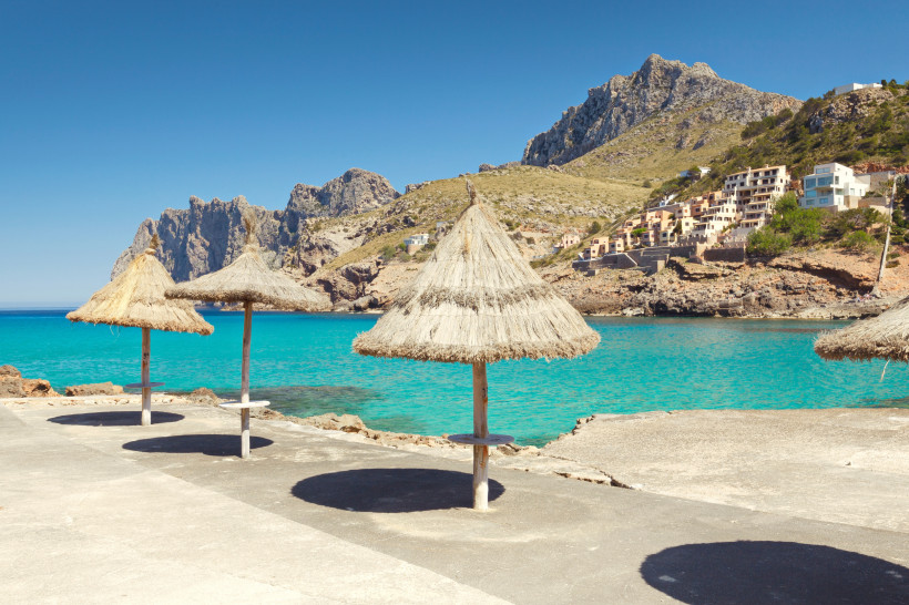 Küstenabschnitt in Cala San Vicente mit Sonnenschirmen, türkisfarbenem Wasser und markanter Bergkulisse im Hintergrund.