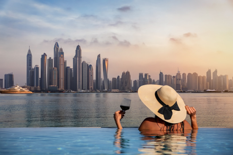 Luxusurlaub, Dubai Eine Frau mit Hut und einem Glas Wein in der Hand steht am Rand eines Infinity Pools und schaut auf die Skyline der Dubai Marina