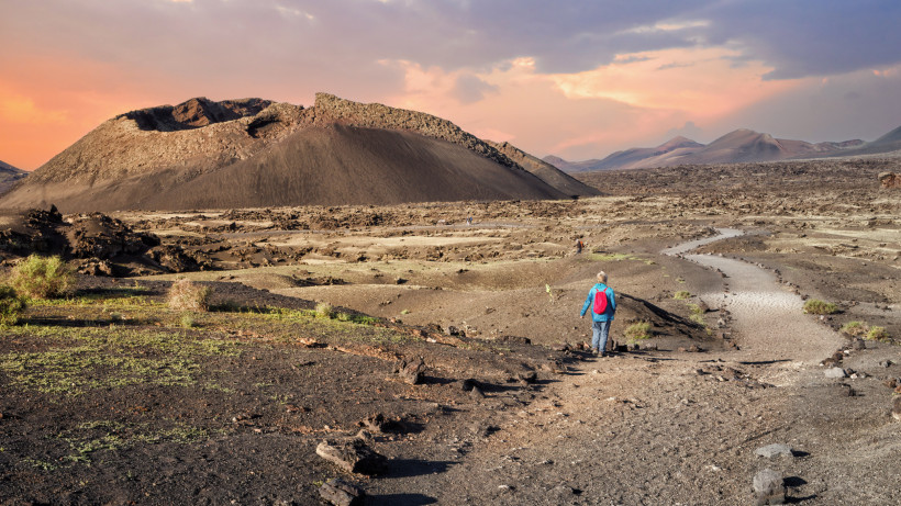 Wanderer auf einem Vulkanpfad im Nationalpark Timanfaya auf Lanzarote, umgeben von schwarzer Lavalandschaft und Kratern bei Sonnenuntergang.