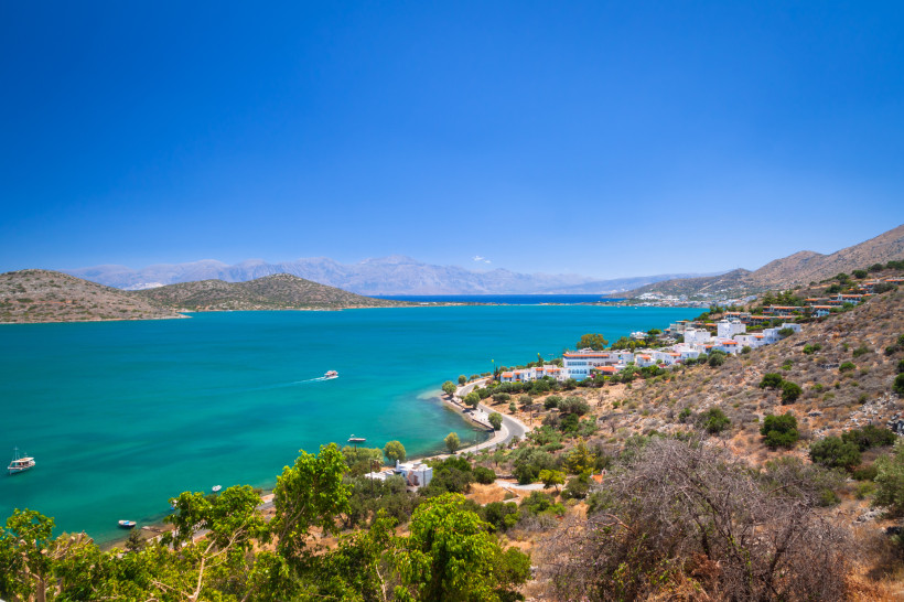 Blick über die Mirabello-Bucht mit türkisblauem Wasser, Küstenstraße und Ortschaft am Hang, dahinter Bergkette