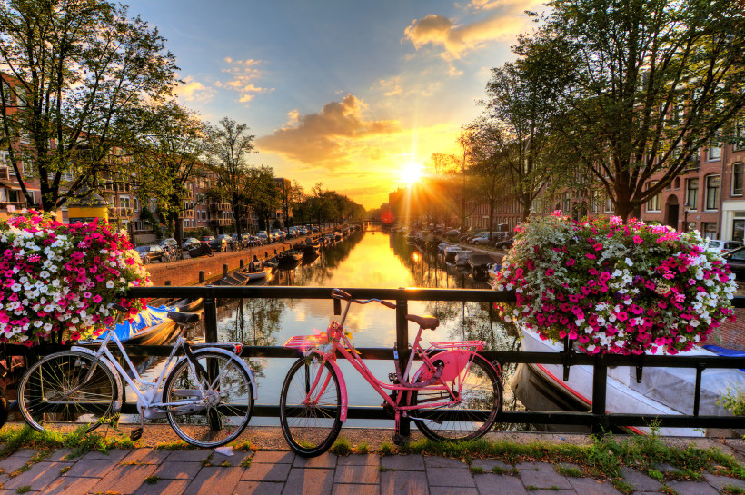 Amsterdam bei Sonnenuntergang: Blick von einer Brücke auf eine ruhige Gracht, links und rechts Baumreihen und Backsteinfassaden. Zwei Fahrräder lehnen am Geländer, daneben üppige Blumenampeln. Goldenes Licht spiegelt sich im Wasser mit Booten.