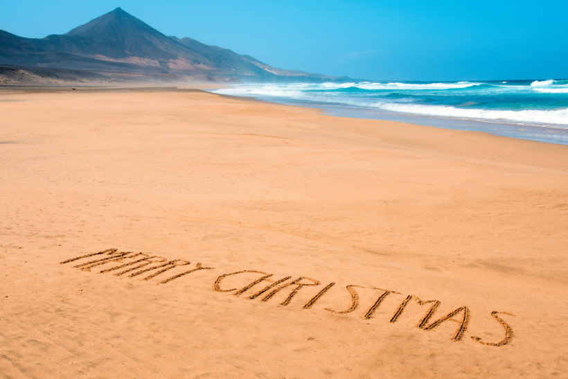 Fuerteventura Weiter Sandstrand mit dem Schriftzug 'Merry Christmas' im Sand. Im Hintergrund Wellen und ein Bergmassiv unter blauem Himmel