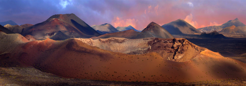Lanzarote Weite Vulkanlandschaft mit rötlich-braunen Kratern und dunklem Lavagestein unter einem farbenprächtigen Himmel bei Sonnenuntergang. Im Hintergrund ragen weitere Vulkankegel auf.
