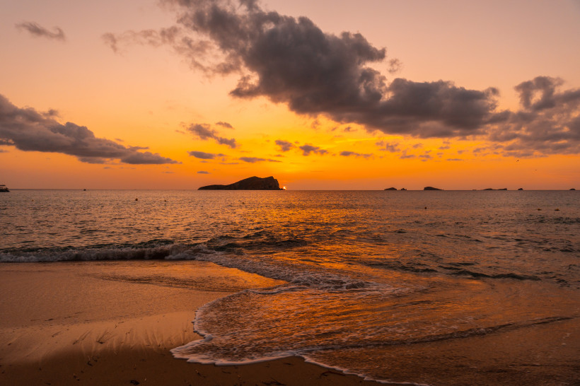 Sonnenuntergang über dem Meer bei Cala Comte mit Sandstrand, sanften Wellen und Inseln am Horizont