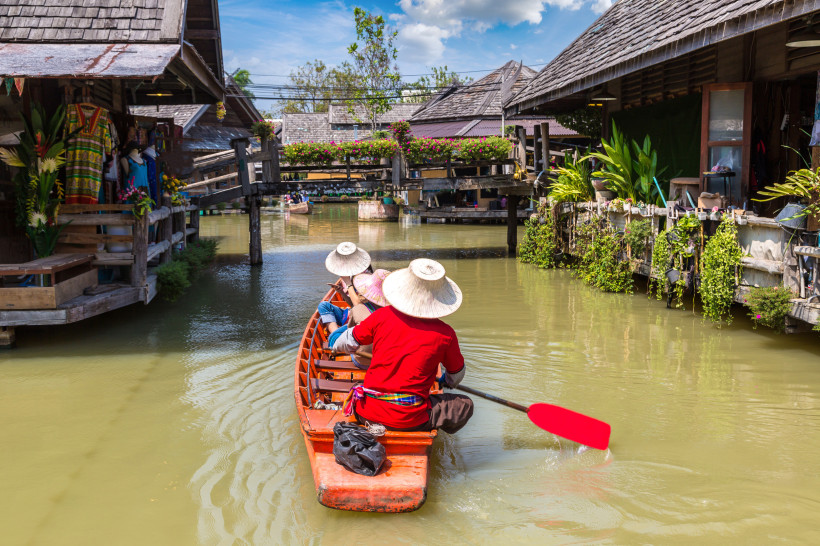 Traditioneller Floating Market in Thailand mit Holzbooten und Kanälen bei Tageslicht Touristen in einem traditionellen Holzboot auf einem Kanal im thailändischen Floating Market, umgeben von Holzhäusern und Pflanzen bei Sonnenschein
