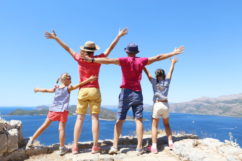 Kroatien Eine Familie mit zwei Kindern steht auf einem Felsvorsprung mit Blick auf das Meer. Alle haben die Arme ausgebreitet und schauen fröhlich in die Ferne. Im Hintergrund sind das tiefblaue Meer, kleine Inseln und Berge zu sehen.