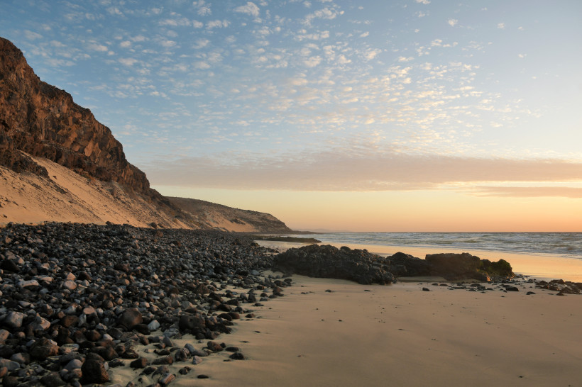 Strandbucht von Esquinz mit Sand, dunklen Steinen und Felsküste am Meer bei Abendlicht