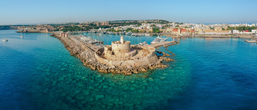 Der Mandraki-Hafen auf Rhodos: Im Vordergrund eine antike Säule mit einer Bronzestatue eines Hirsches, Symbol der Insel. Ein Angler sitzt am Rand des Kais. Im Hintergrund liegt die mittelalterliche Festung Agios Nikolaos, umgeben von Segelbooten und türki